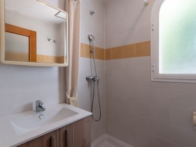 Salle de bain fonctionnelle avec meuble-lavabo en bois clair, douche et fenêtre arquée dépolie, agrémentée de carreaux blancs et d'une bande décorative ocre.