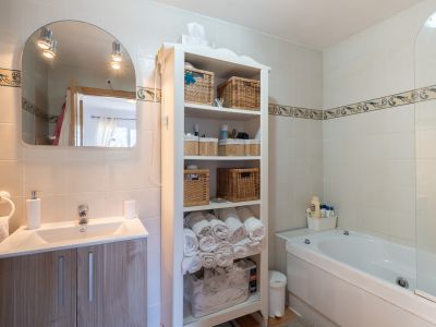 Salle de bain moderne avec meuble-lavabo imitation bois clair, miroir arqué éclairé et baignoire. Comprend une étagère blanche garnie de paniers en osier et des murs carrelés avec frise décorative.