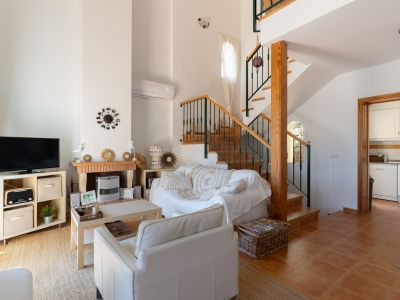 Salon chaleureux avec cheminée en brique et bois, canapé blanc et escalier en bois avec rampe verte, menant à l'étage supérieur et à la cuisine.