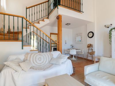 Salon lumineux avec canapé blanc, table basse en bois clair et un escalier en bois distinctif avec balustrade en métal vert foncé. Un coin repas est intégré.