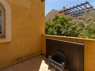 Terrasse extérieure avec murs en stuc ocre texturé et carrelage au sol. Elle dispose d'une fenêtre arquée, d'une jardinière décorative en métal et de vues sur la verdure et une pergola.