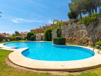 Vaste piscine commune de forme irrégulière sur une pelouse verdoyante, avec des bâtiments résidentiels en terrasses et un mur de roche naturelle en arrière-plan sous un ciel clair.