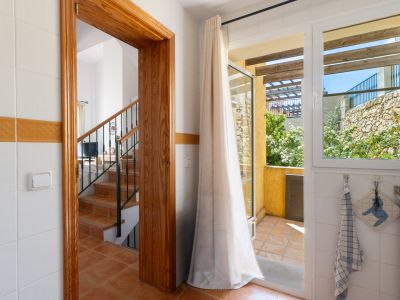 Vue intérieure carrelée, présentant une porte en bois vers un escalier en terre cuite et une porte vitrée menant à une terrasse extérieure avec mur de pierre et verdure.