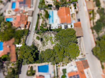 Vue aérienne d'une parcelle non bâtie avec des arbres, bordée par des routes et des propriétés résidentielles avec piscines et toits rouges.