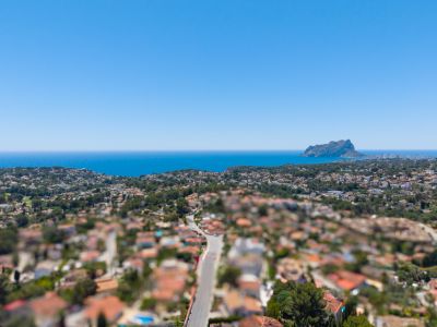 Vue aérienne panoramique de la côte de Benissa, avec la mer Méditerranée, le Peñón de Ifach et un quartier résidentiel verdoyant sous un ciel bleu dégagé.