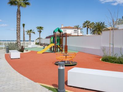 Aire de jeux moderne avec revêtement de sécurité rouge, structure de jeux colorée et carrousel, entourée de palmiers et vue lointaine sur la mer.