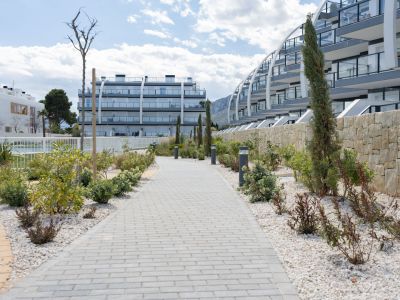 Allée pavée bordée de gravier blanc, plantes méditerranéennes et bornes lumineuses, menant vers des appartements modernes aux balcons vitrés et un mur de pierre naturelle.