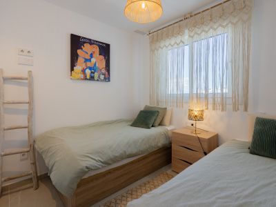 Chambre avec deux lits simples à cadres en bois clair et literie verte. Rideaux en macramé, lampe de table à abat-jour décoré et tableau pop art coloré agrémentent l'espace.