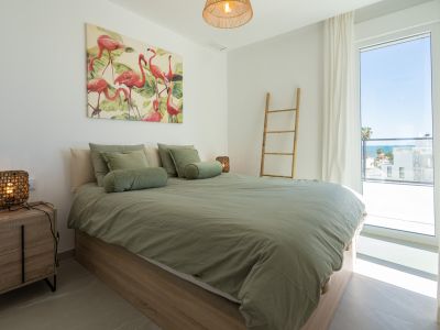 Chambre lumineuse avec lit en bois clair, literie vert sauge et tableau de flamants roses. Grande fenêtre offrant une vue partielle sur la mer.