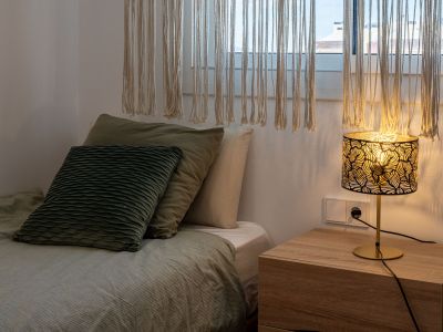 Coin chambre apaisant avec un lit aux parures vertes, une table de chevet en bois clair et une lampe décorative à abat-jour ajouré. Des rideaux en macramé tamisent la lumière naturelle.