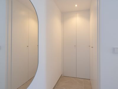 Couloir moderne avec placards intégrés blancs aux poignées discrètes. Un miroir ovale reflète l'éclairage encastré chaleureux sur le sol carrelé.