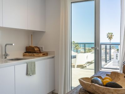 Cuisine blanche moderne avec des armoires épurées et un évier, baignée de lumière naturelle, donnant directement sur une terrasse avec vue imprenable sur la mer et palmiers.