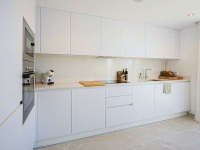 Cuisine moderne et fonctionnelle avec armoires blanches sans poignées, plan de travail clair et appareils intégrés. Four, micro-ondes, plaque à induction, éclairage encastré et sol carrelé clair.