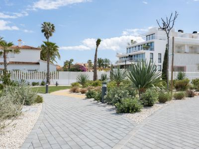 Des allées pavées bordées de gravier blanc traversent un aménagement paysager méditerranéen luxuriant, avec palmiers et agaves, menant vers des immeubles modernes et une maison traditionnelle sous un ciel bleu.