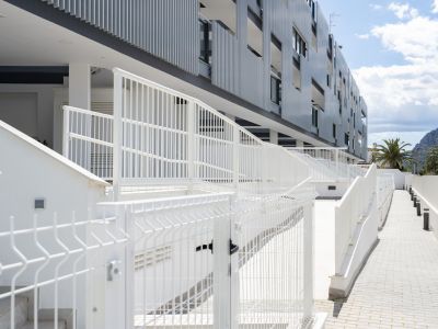 Extérieur moderne d'un immeuble d'appartements avec balustrades blanches, allées pavées et escaliers, avec palmiers et montagnes en arrière-plan sous un ciel bleu.