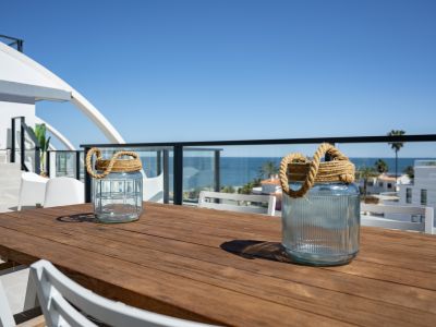 Grande terrasse extérieure avec table en bois et chaises blanches, offrant des vues partielles sur la mer Méditerranée et le paysage côtier.
