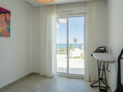 Pièce lumineuse avec sol carrelé et porte coulissante vitrée menant à une terrasse offrant vue sur mer et palmiers. Une planche à repasser et une peinture colorée sont visibles.