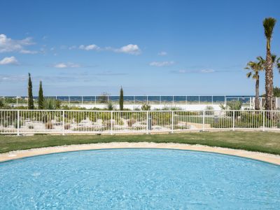Piscine commune avec bord incurvé, pelouse verte et jardin méditerranéen. Offre une vue dégagée sur la mer sous un ciel bleu.