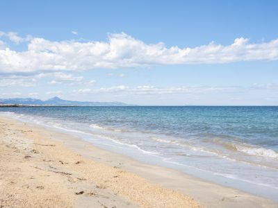 Plage de sable doré avec de douces vagues atteignant le rivage, menant à des eaux turquoise claires. Des montagnes lointaines et un brise-lames encadrent l'horizon sous un ciel nuageux.