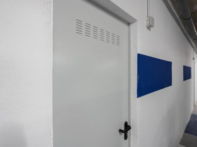 Porte de débarras métallique blanche avec fentes d'aération et poignée noire, dans un couloir de garage aux bandes murales bleues.