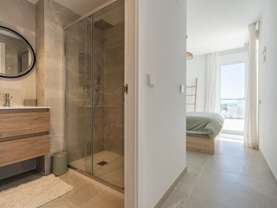 Salle de bain moderne avec carrelage effet marbre, meuble vasque en bois et douche vitrée. La chambre adjacente offre une vue mer lointaine.