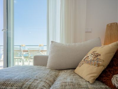 Salon confortable avec canapé texturé et coussins, s'ouvrant sur une terrasse extérieure meublée sous un ciel bleu. Lumière naturelle et lampe en corde.
