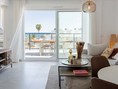 Salon lumineux avec carrelage clair et accès direct à une terrasse offrant une vue panoramique sur la mer et un palmier. Présente un luminaire suspendu moderne et des rideaux blancs.