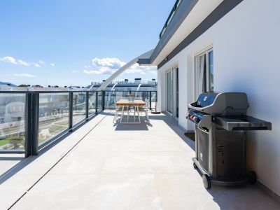 Spacieuse terrasse avec carrelage clair, balustrades en verre et barbecue moderne. Comprend un coin repas extérieur et des vues sur les bâtiments et les montagnes.