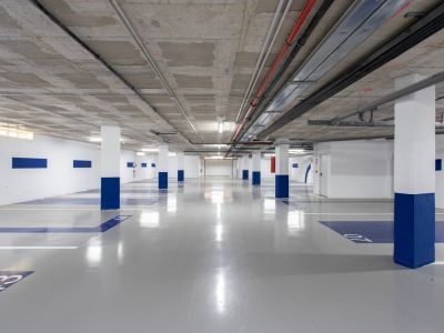 Vaste parking souterrain avec plafond en béton brut et conduits industriels apparents. Piliers blancs à base bleue, sol en résine grise avec marquages numérotés et éclairage fluorescent. Borne de recharge pour VE.