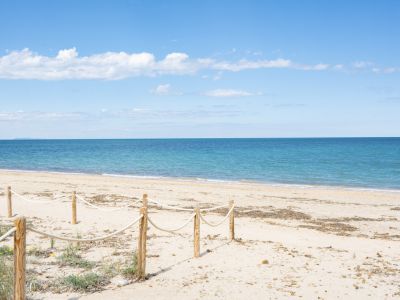 Vaste plage de sable aux eaux turquoise calmes sous un ciel bleu clair, avec une clôture en corde au premier plan et une terre lointaine à l'horizon.