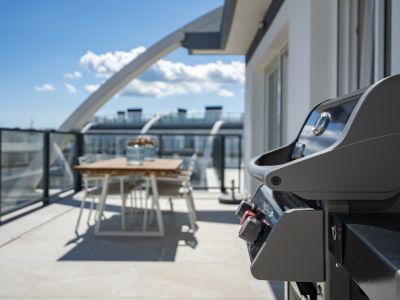 Vaste terrasse sur le toit avec un barbecue à gaz moderne et un ensemble de salle à manger extérieur en bois, balustrade en verre et carrelage clair sous un ciel dégagé.