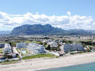 Vue aérienne d'appartements blancs modernes bordant une plage de sable doré et la mer turquoise, avec l'imposante chaîne du Montgó en arrière-plan.