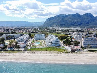 Vue aérienne de complexes résidentiels modernes avec piscines et palmiers, situés en front de mer sur une plage de sable, avec l'imposante montagne de Dénia en arrière-plan.