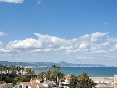 Vue panoramique de la côte méditerranéenne avec des bâtiments résidentiels, des palmiers, une plage de sable et des montagnes lointaines sous un ciel bleu nuageux.