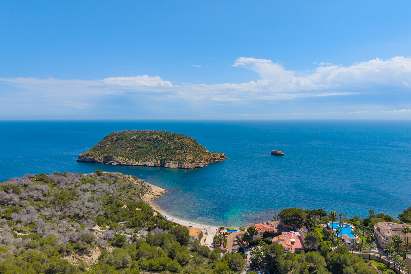 Vue aérienne de la côte de Jávea avec l'Isla del Descubridor, la plage et une zone résidentielle avec piscine.