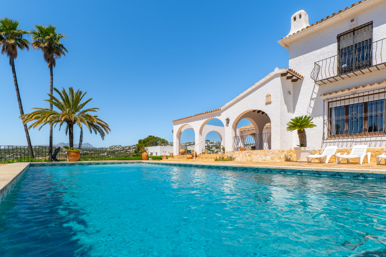 Piscine turquoise avec bordure en mosaïque et villa blanche de style méditerranéen avec arcades.