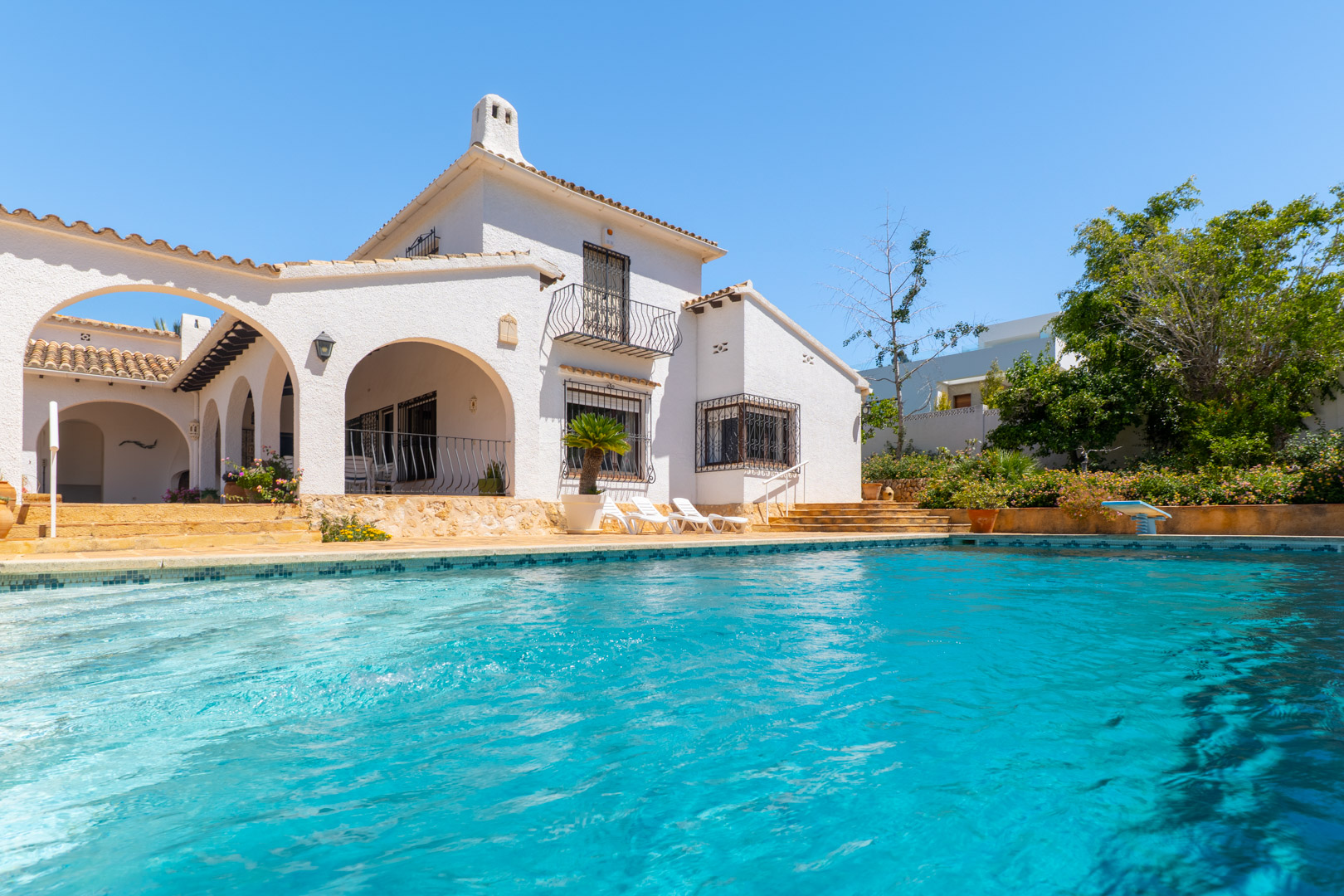 Piscine turquoise bordée de mosaïque devant la façade blanche.