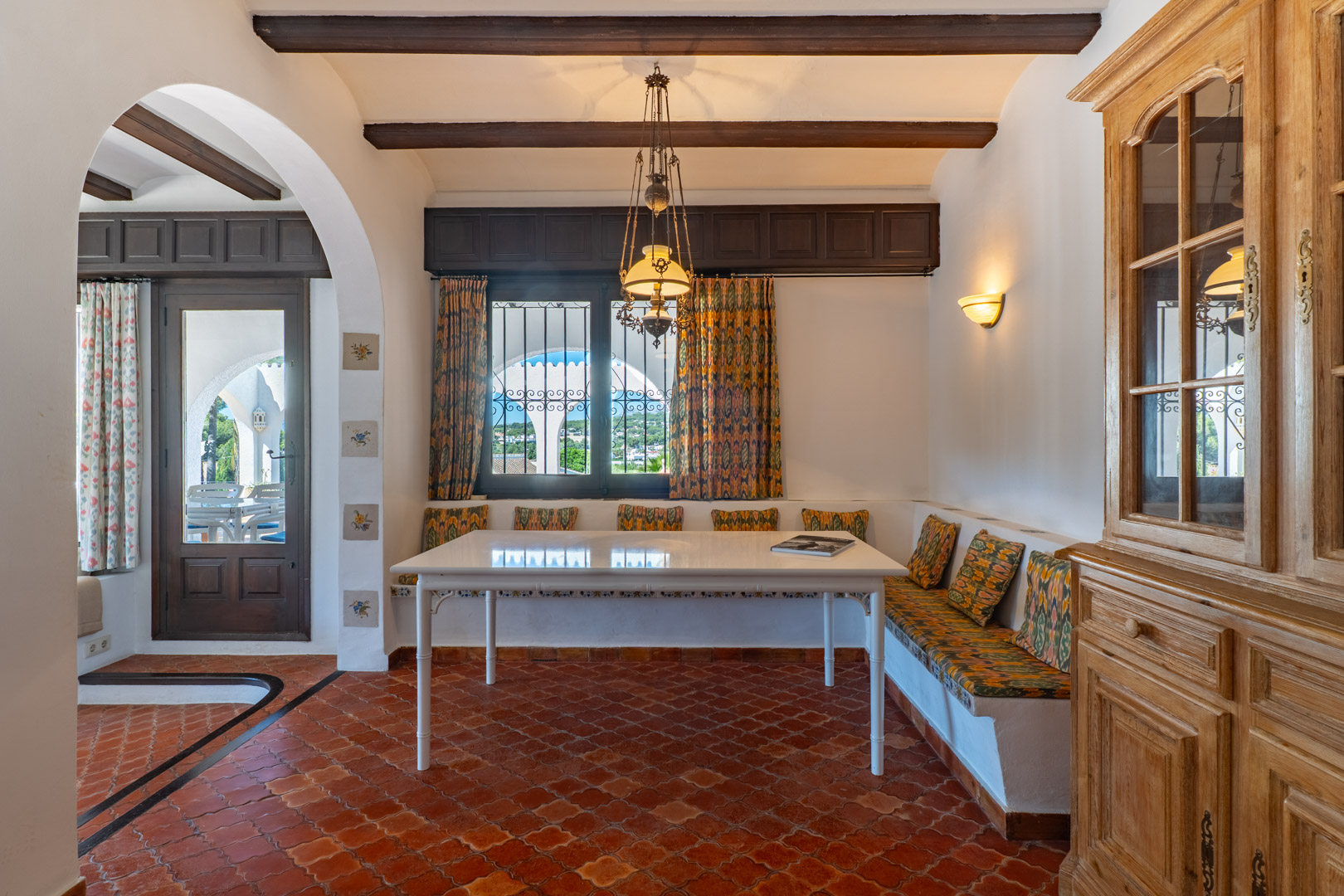 Salle à manger avec banquette intégrée, table blanche et sol en terre cuite, éclairée par un lustre.