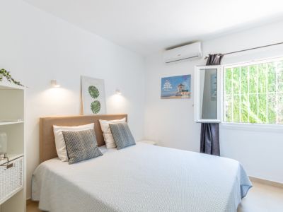 Chambre avec lit double, tête de lit en tissu beige, climatisation et fenêtre protégée par une grille, offrant une vue sur la verdure.