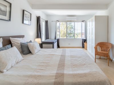 Chambre spacieuse avec grand lit, carrelage clair, armoire blanche et fenêtre sécurisée offrant une vue sur la verdure extérieure, avec climatisation.