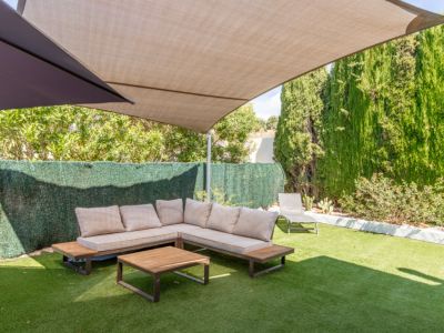 Espace lounge extérieur ombragé avec canapé en L aux coussins clairs et table basse en bois sur gazon artificiel. De hautes haies et arbres offrent intimité sous une voile d'ombrage beige.