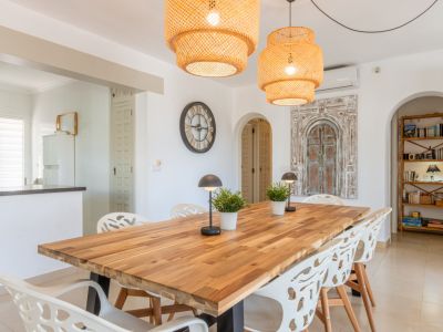 Salle à manger lumineuse avec une grande table en bois, des chaises blanches contemporaines et deux suspensions en osier. Des arches mènent à la cuisine et à une bibliothèque.