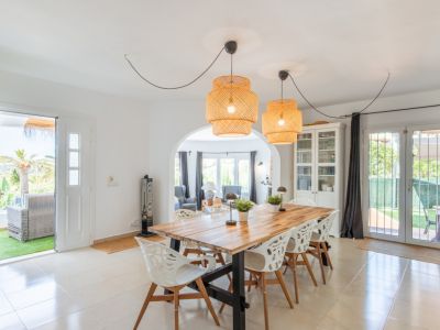 Salle à manger lumineuse avec une grande table en bois, des suspensions en rotin et un sol carrelé. Elle donne directement sur une terrasse privée et s'ouvre sur un salon par une arche.