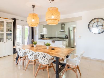 Salle à manger lumineuse avec une grande table en bois, huit chaises blanches design et deux suspensions tressées. Elle s'ouvre sur une cuisine moderne équipée d'appareils en acier inoxydable.