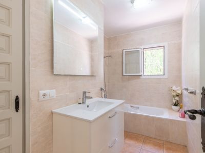 Salle de bain avec carrelage beige clair, meuble-lavabo blanc et grand miroir éclairé. Comprend une baignoire et une fenêtre apportant la lumière naturelle.