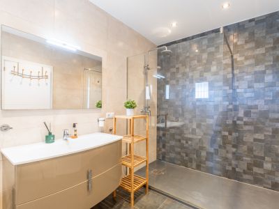Salle de bain moderne avec meuble-lavabo beige laqué, miroir éclairé et douche à l'italienne avec paroi vitrée et carrelage mosaïque gris foncé. Murs clairs et sol sombre.