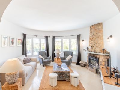 Salon lumineux avec mur incurvé de grandes fenêtres, cheminée imposante revêtue de pierre naturelle et carrelage clair. Un tapis en jute délimite l'espace.