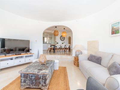 Salon spacieux avec carrelage clair, canapé confortable et table basse rustique en bois sur tapis de jute. Une arche mène à la salle à manger surélevée avec des sièges modernes et des suspensions en rotin.