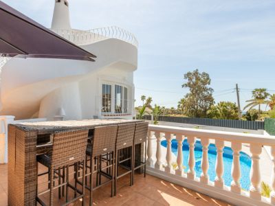 Terrasse extérieure carrelée avec un bar en rotin et des tabourets, offrant une vue sur la piscine privée. La villa blanche présente un détail architectural spiralé distinctif.
