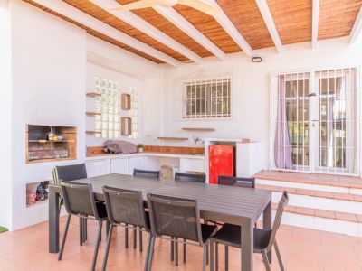 Terrasse extérieure couverte avec barbecue intégré, cuisine d'été fonctionnelle avec mini-réfrigérateur rouge et grande table à manger. Le sol est en terre cuite et le plafond est latté.